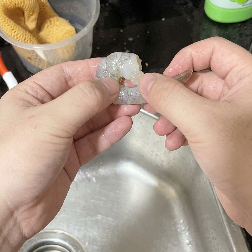 Deux mains décortiquant soigneusement une crevette grise crue au-dessus dun évier en inox.