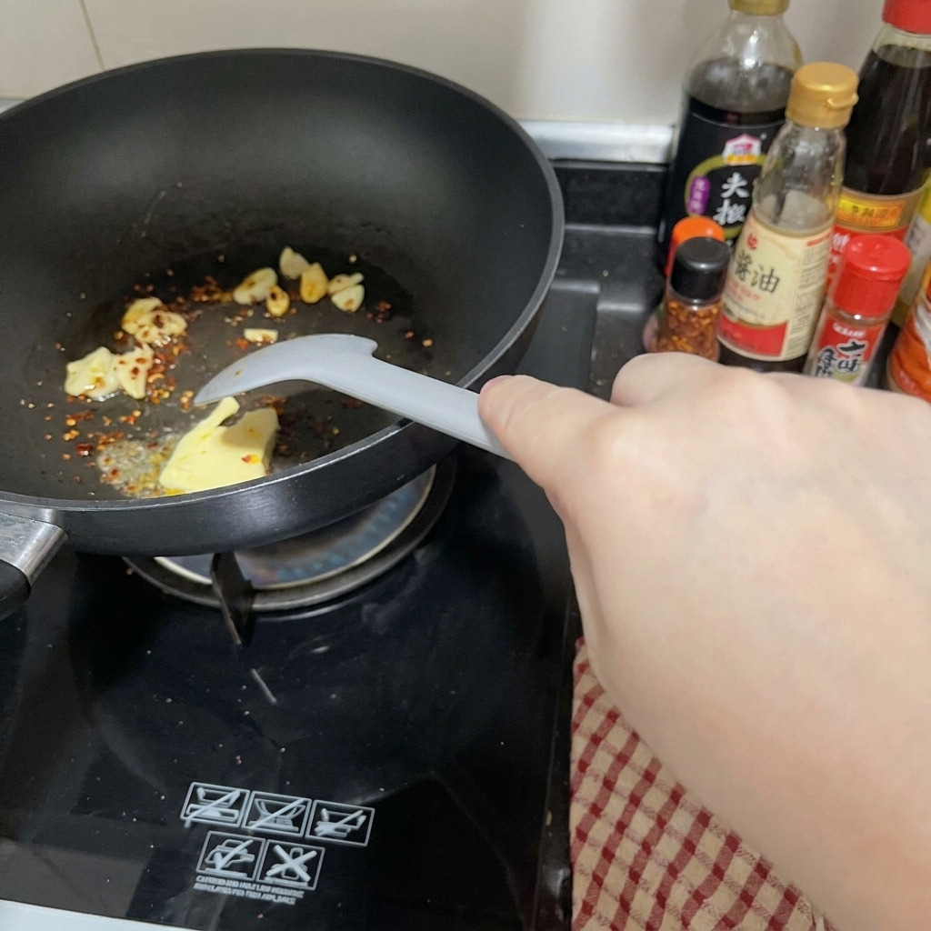Un morceau de beurre fondant dans une poêle noire avec lail et le piment, mélangé par une spatule grise.