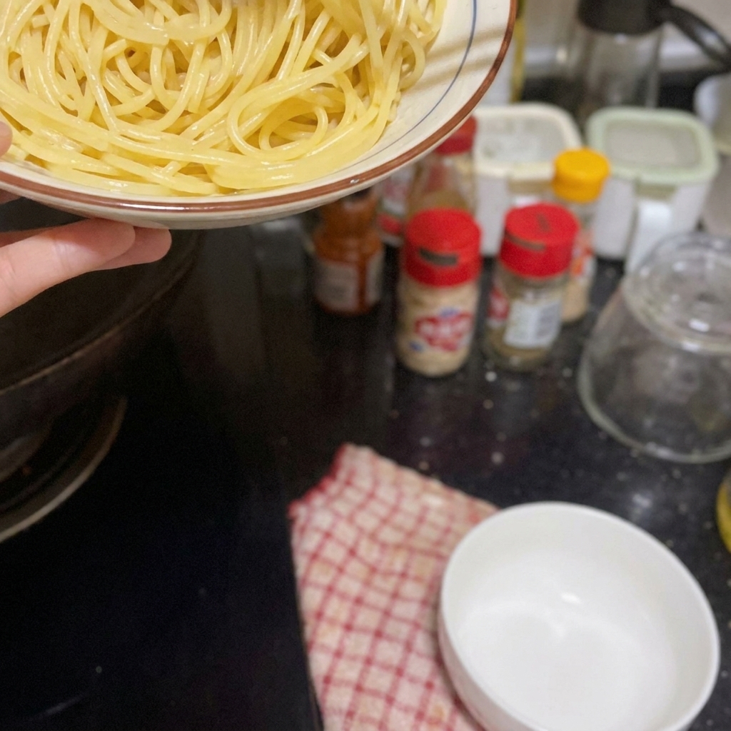 Une main tenant un bol de spaghetti cuits au-dessus dune plaque de cuisson, sapprêtant à les verser dans la poêle.