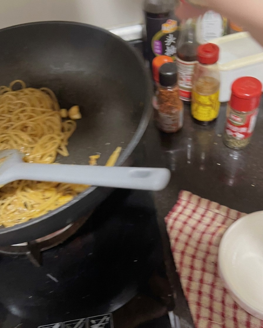 Une main secouant un pot dépices sur une poêle de spaghetti cuits mélangés à lail.
