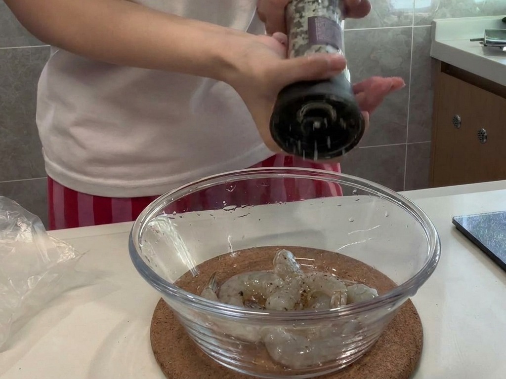 Poivre noir frais moulu au-dessus dun bol en verre contenant des crevettes crues décortiquées.