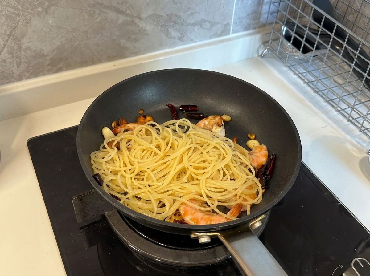 Des spaghettis cuits ajoutés à une poêle remplie de crevettes cuites, dail et de piments séchés.