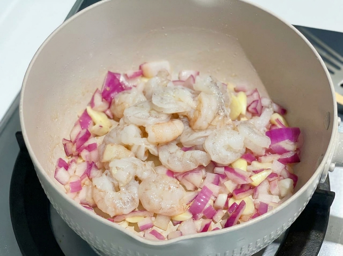 Crevettes crues et déveinées ajoutées dans une poêle avec des oignons rouges sautés.