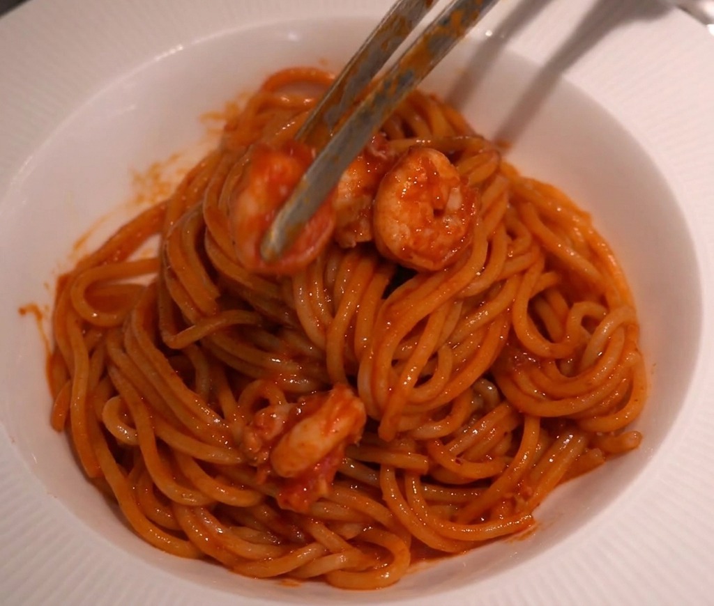 Pince de cuisine plaçant soigneusement des crevettes en sauce sur un dôme de spaghetti.