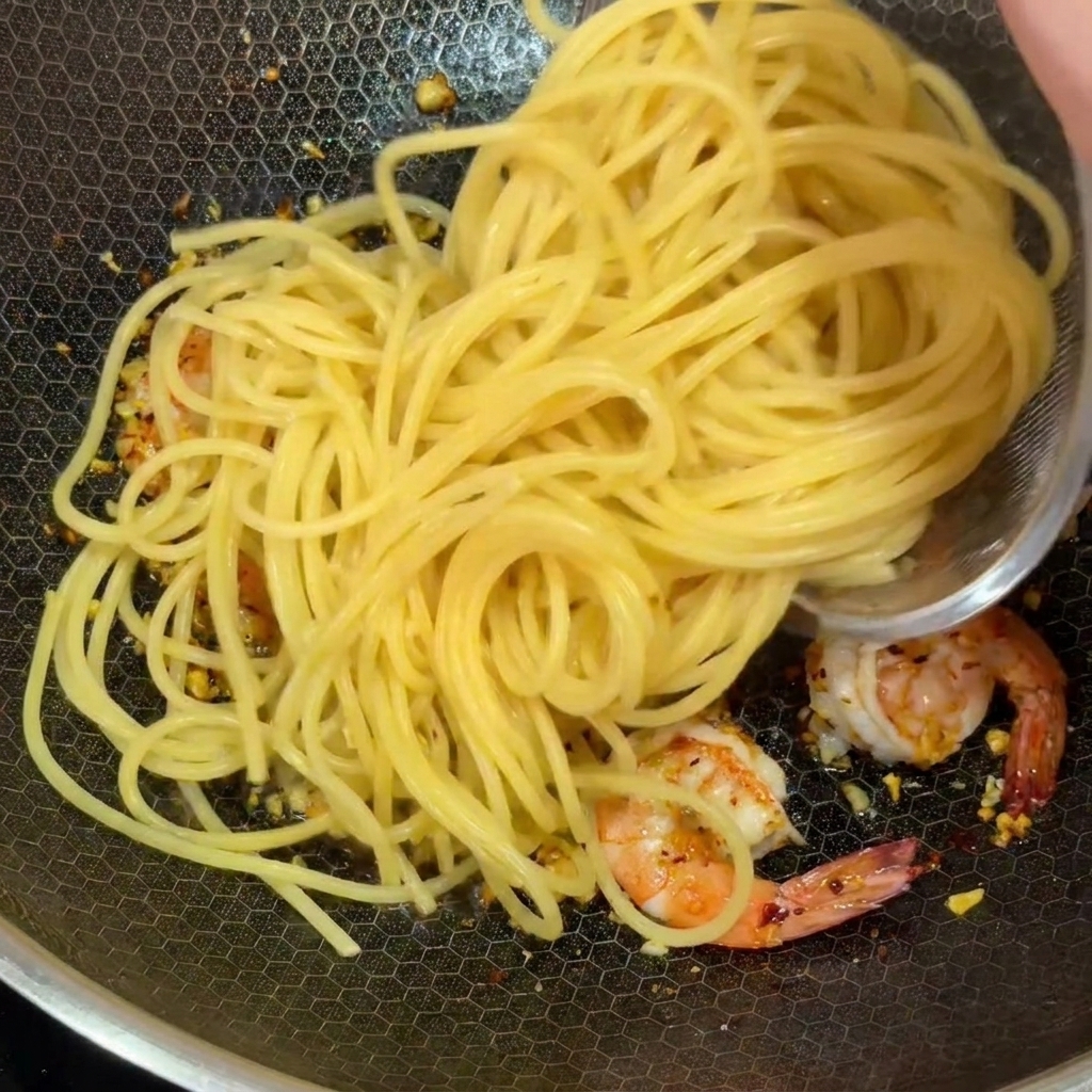 Spaghettis cuits ajoutés dans un wok contenant des crevettes roses et de lail haché.
