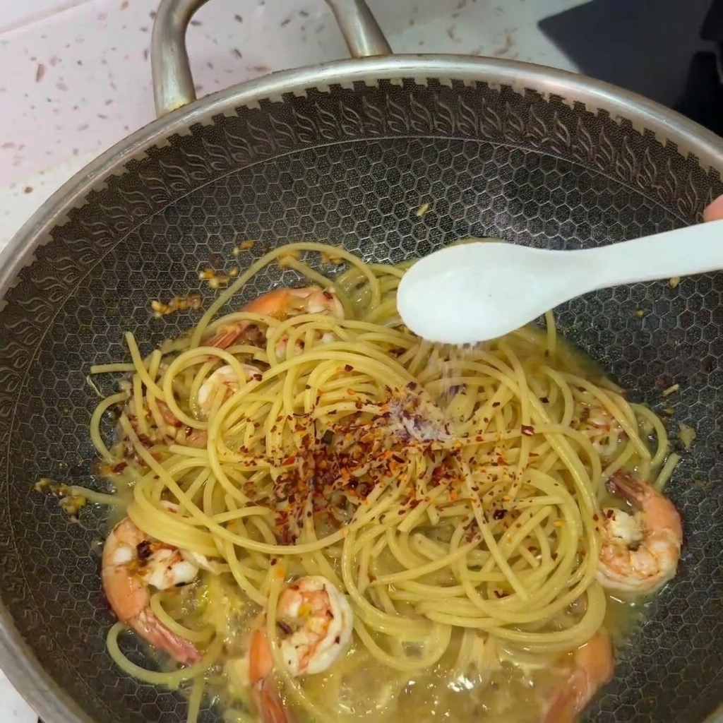 Une cuillère blanche saupoudrant du sel sur les spaghettis et les crevettes, surmontés de piment.