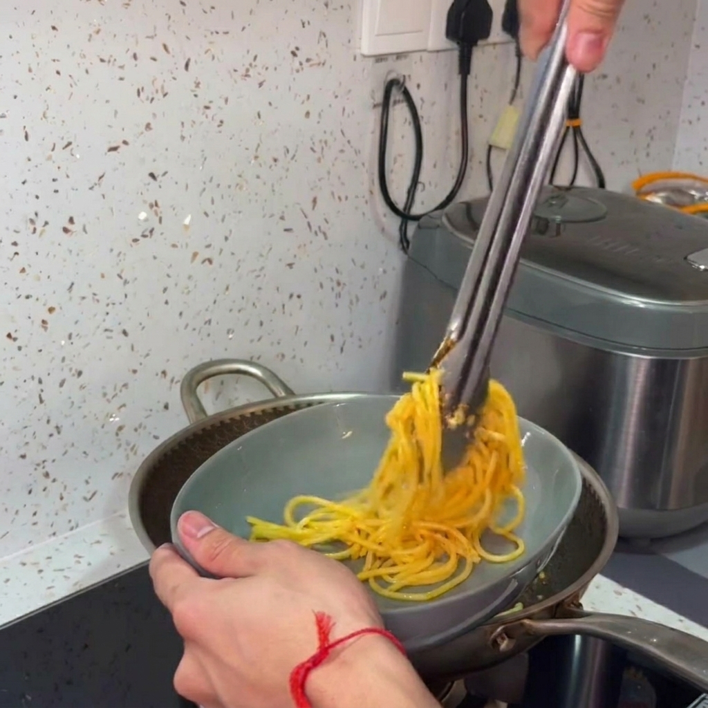 Mains utilisant une pince pour transférer et enrouler les spaghettis dans un bol de service gris.