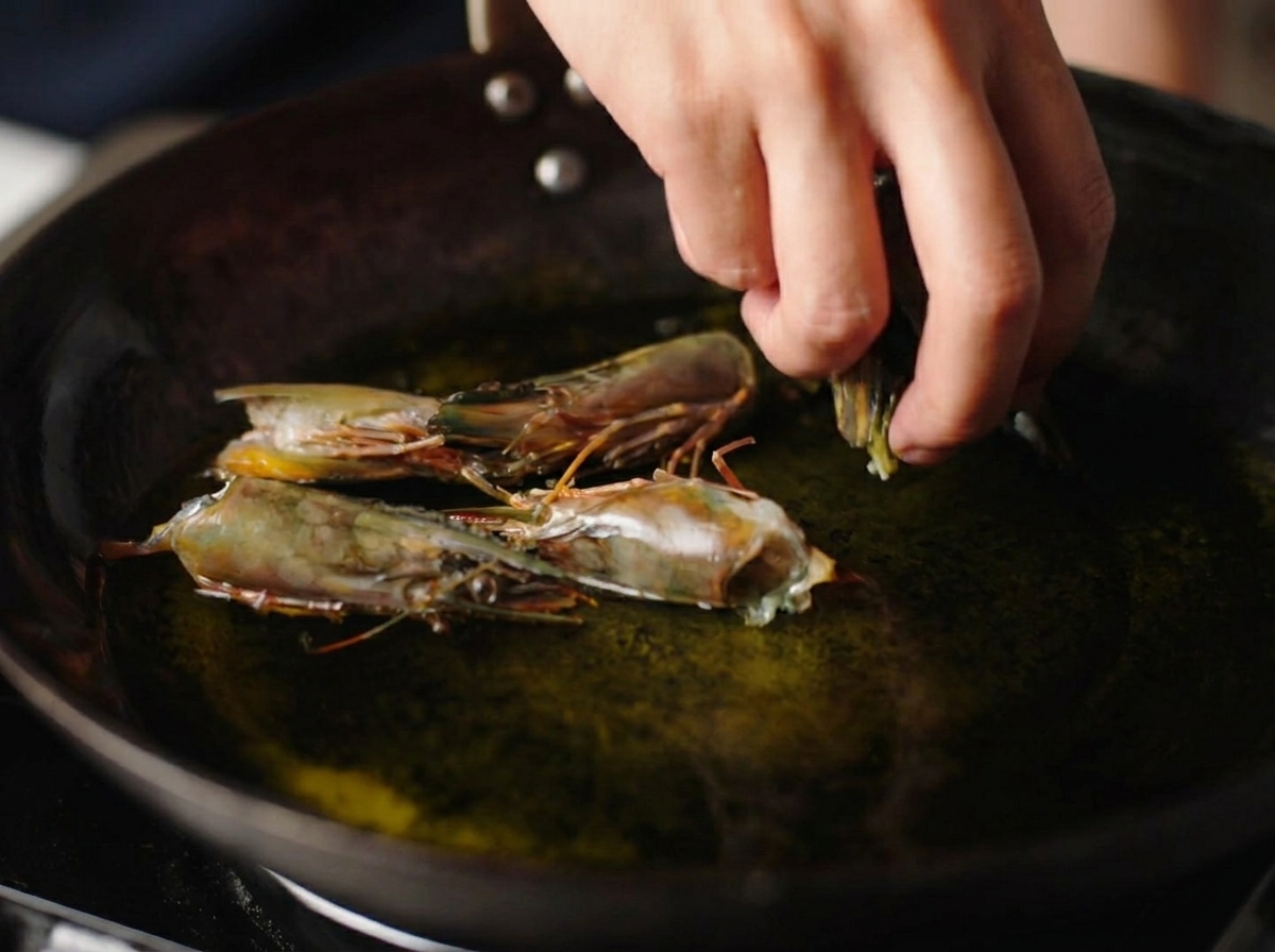 Une main ajoutant des têtes de crevettes crues dans un wok rempli dhuile chaude frémissante.