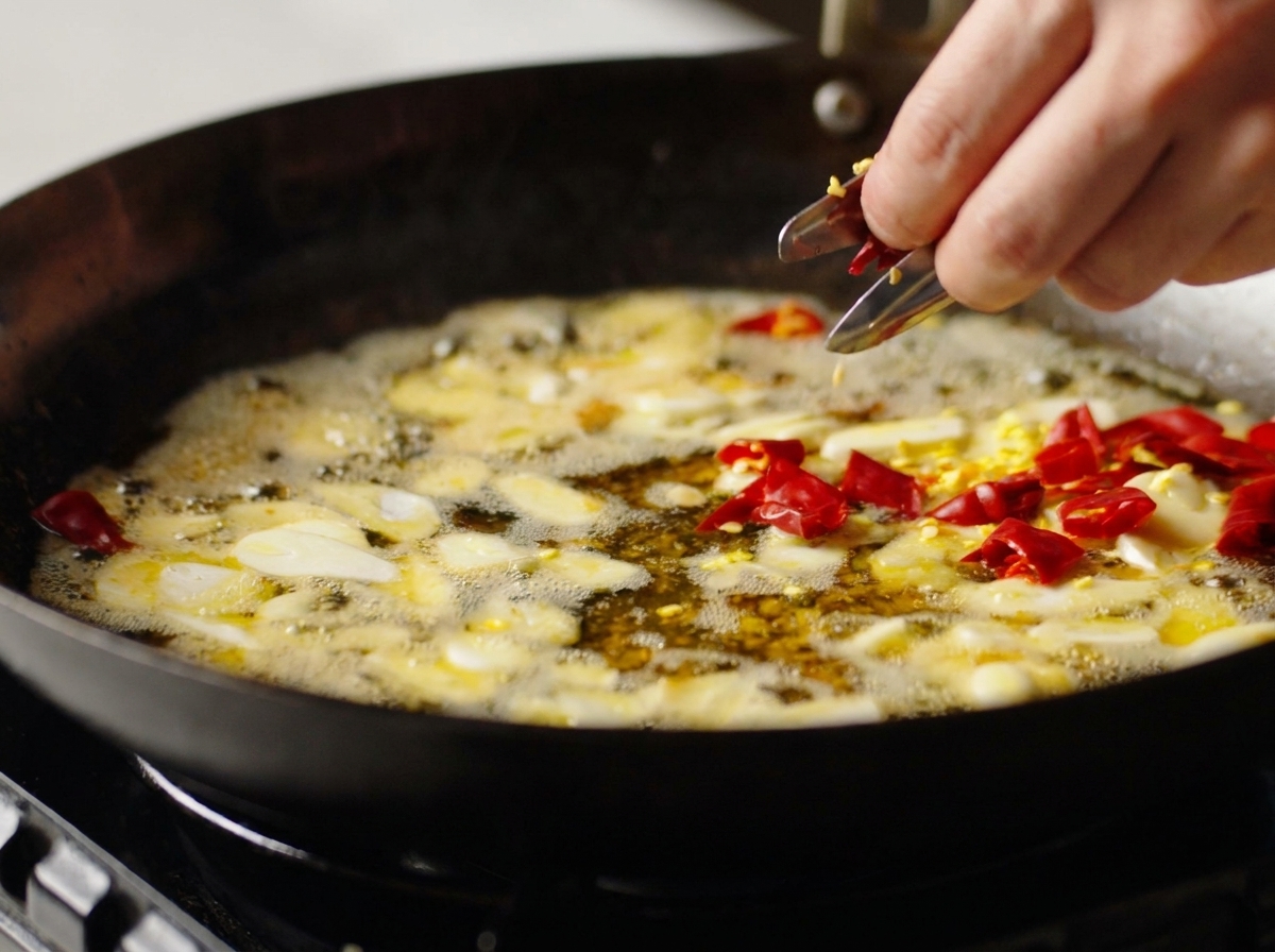 Une main ajoutant des morceaux de piment rouge séché dans un wok avec de lail et de lhuile parfumée.
