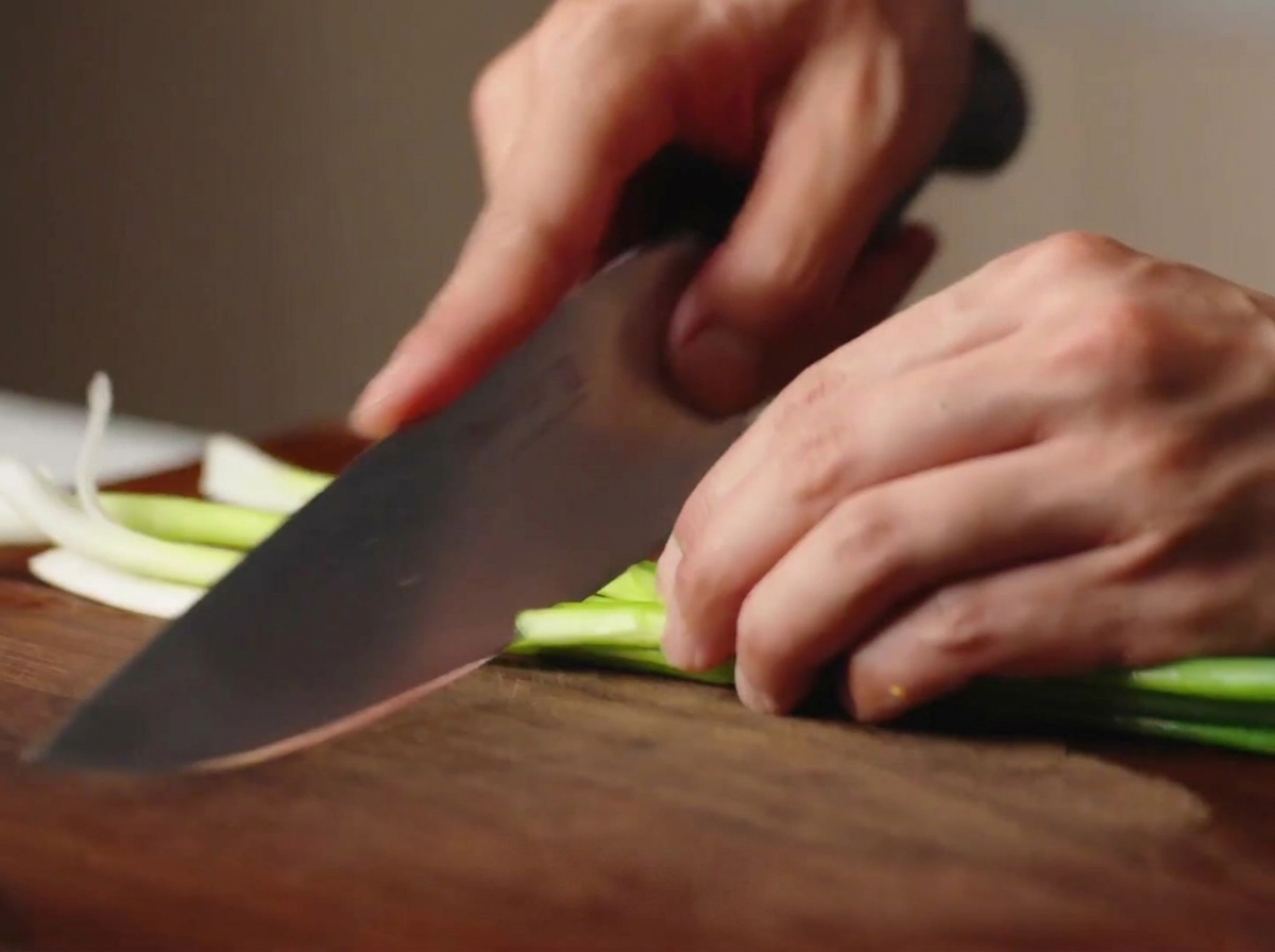 Des mains utilisant un couteau de chef pour couper des oignons verts frais sur une planche en bois.