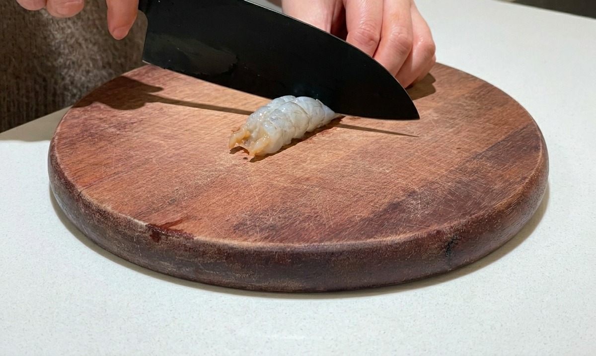 Un couteau incisant le dos dune crevette crue décortiquée sur une planche à découper en bois ronde.