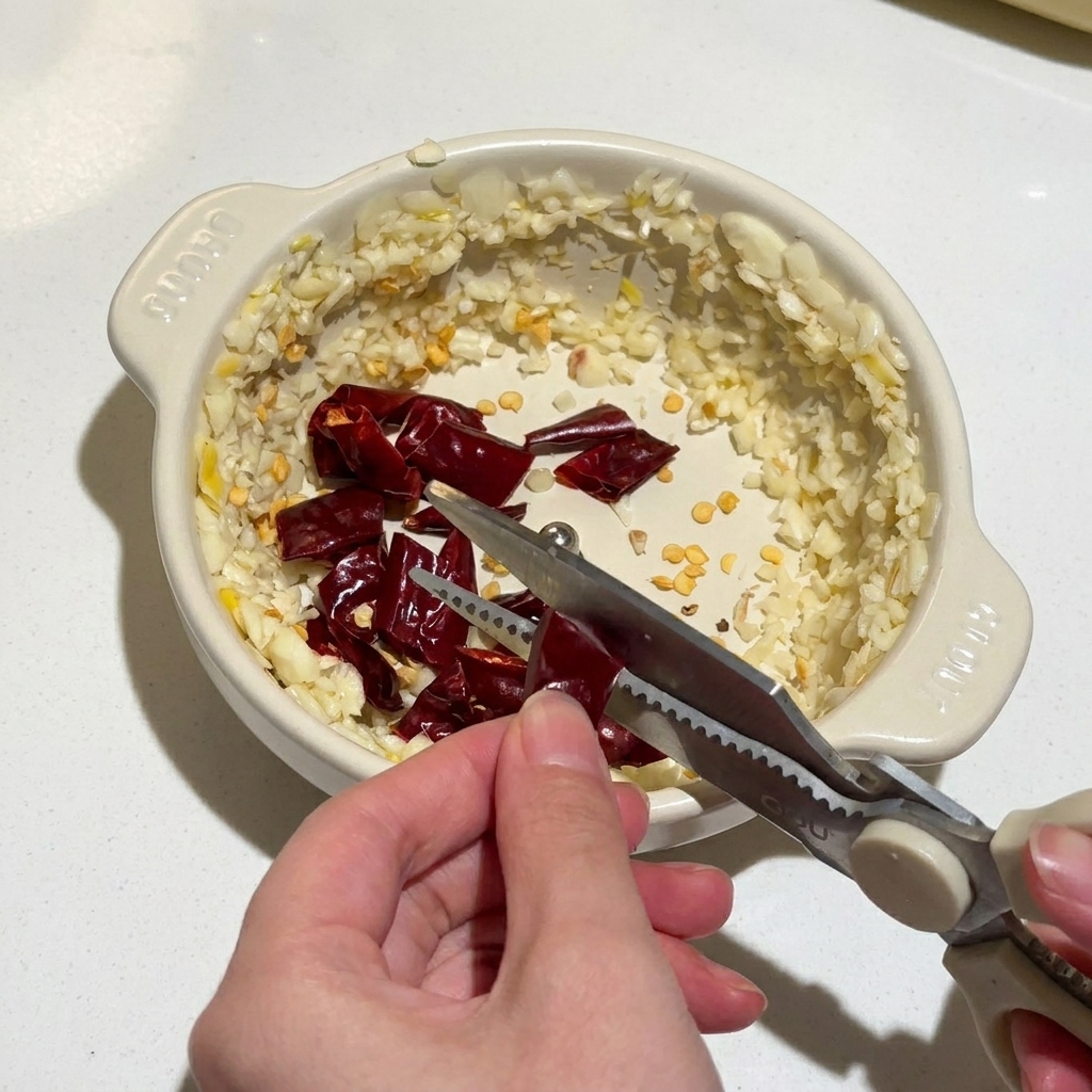 Mains utilisant des ciseaux de cuisine pour couper des piments rouges séchés dans un bol contenant lail haché.