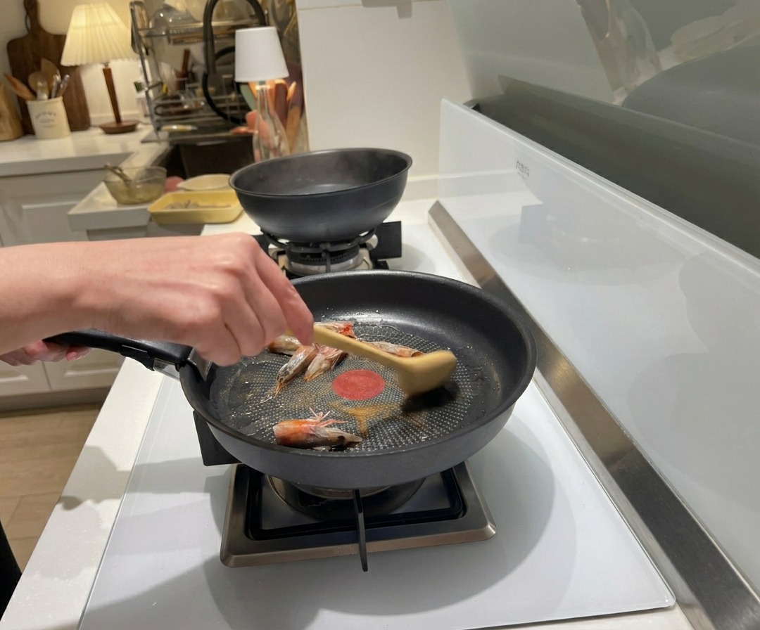Une main utilisant une cuillère en bois pour faire sauter des têtes de crevettes crues dans de lhuile chaude dans une poêle noire.