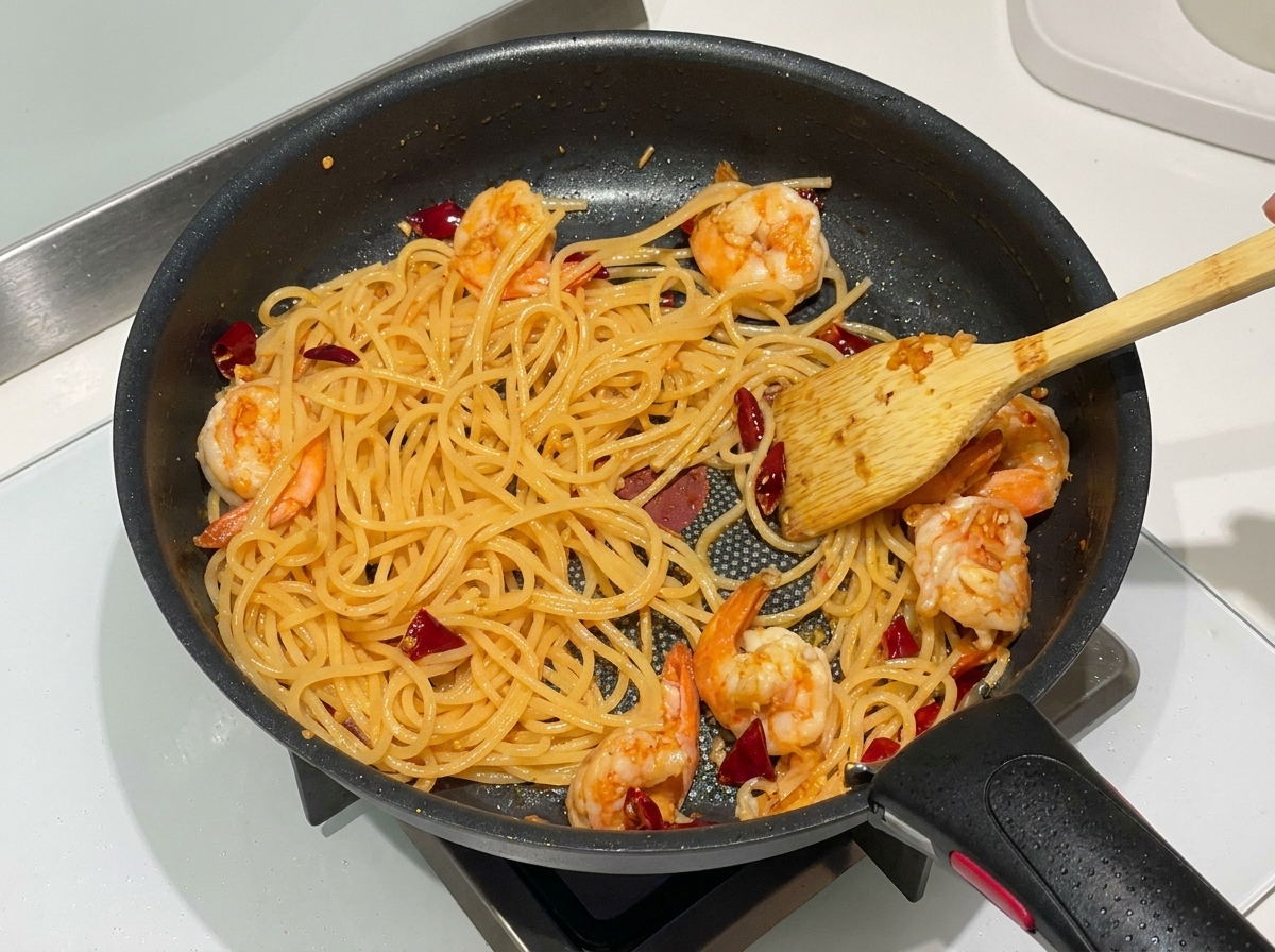 Spaghettis cuits, crevettes roses et piments rouges sautés ensemble dans une poêle antiadhésive noire avec une spatule en bois.