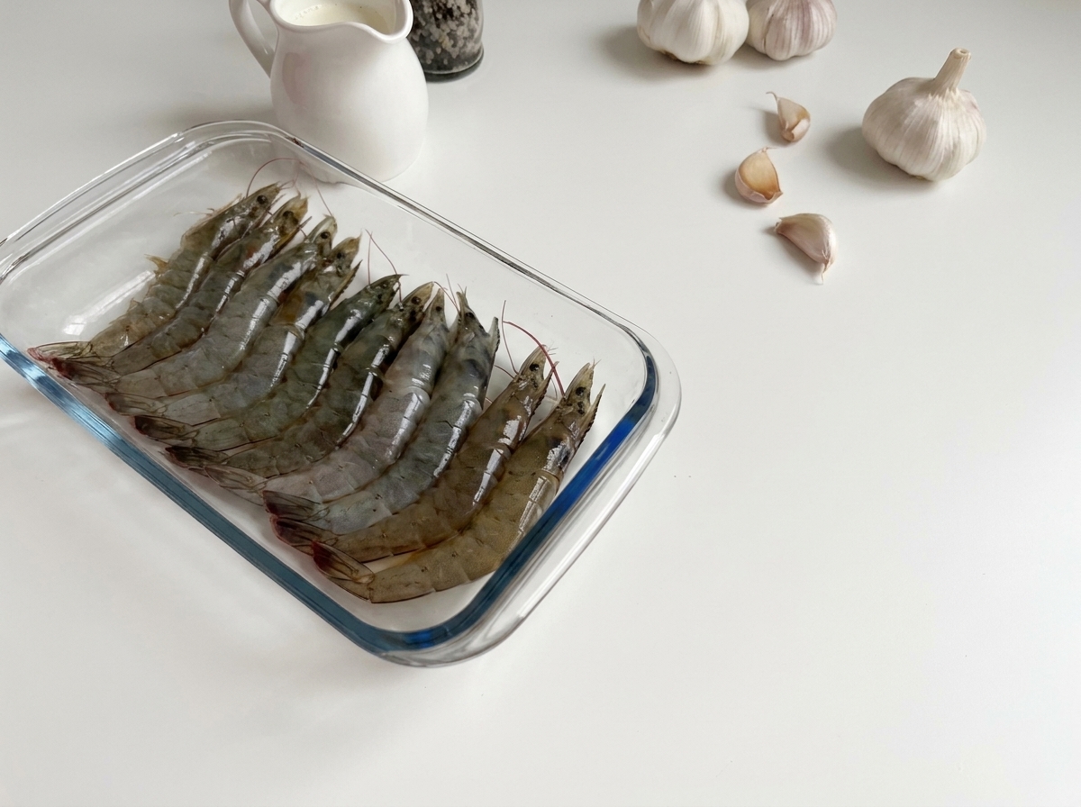 Crevettes grises crues et translucides alignées dans un plat rectangulaire en verre.