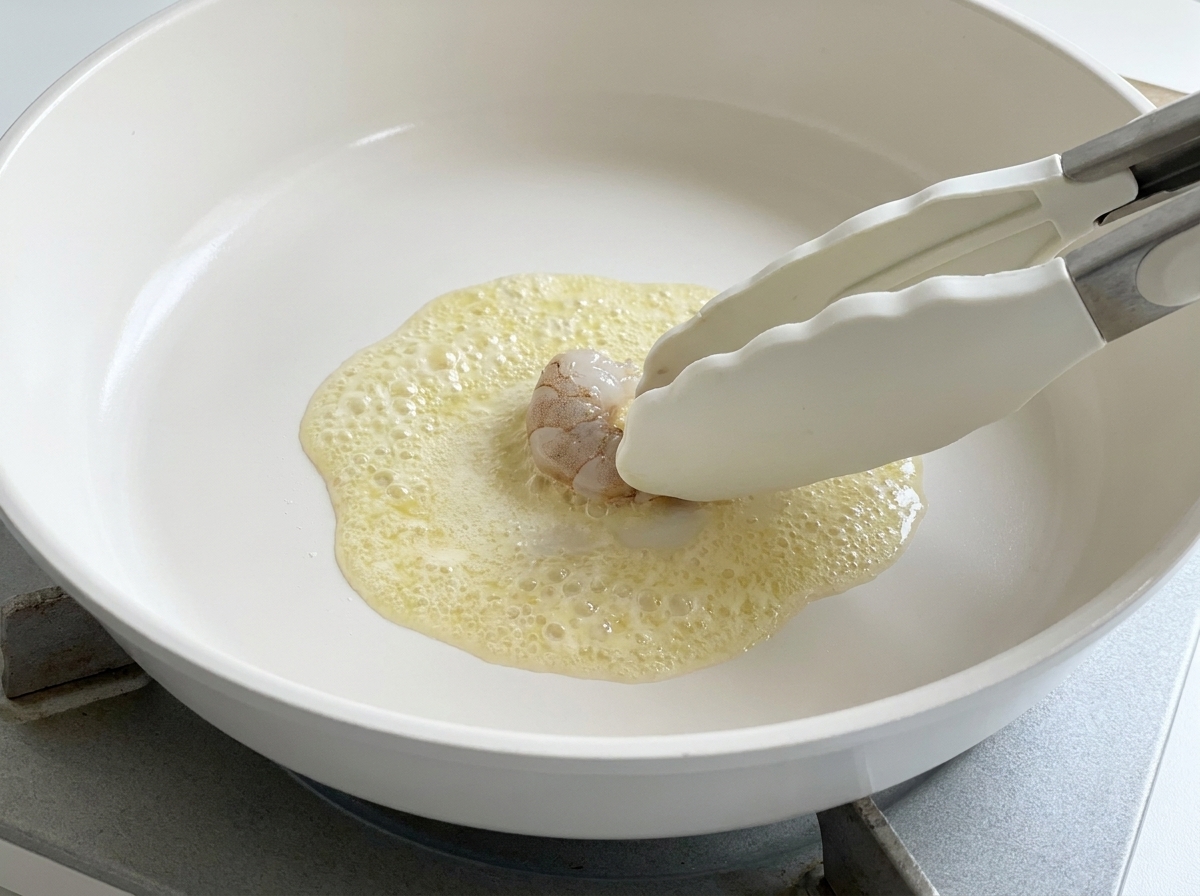 Une pince déposant une crevette crue dans une poêle de beurre fondu frémissant.