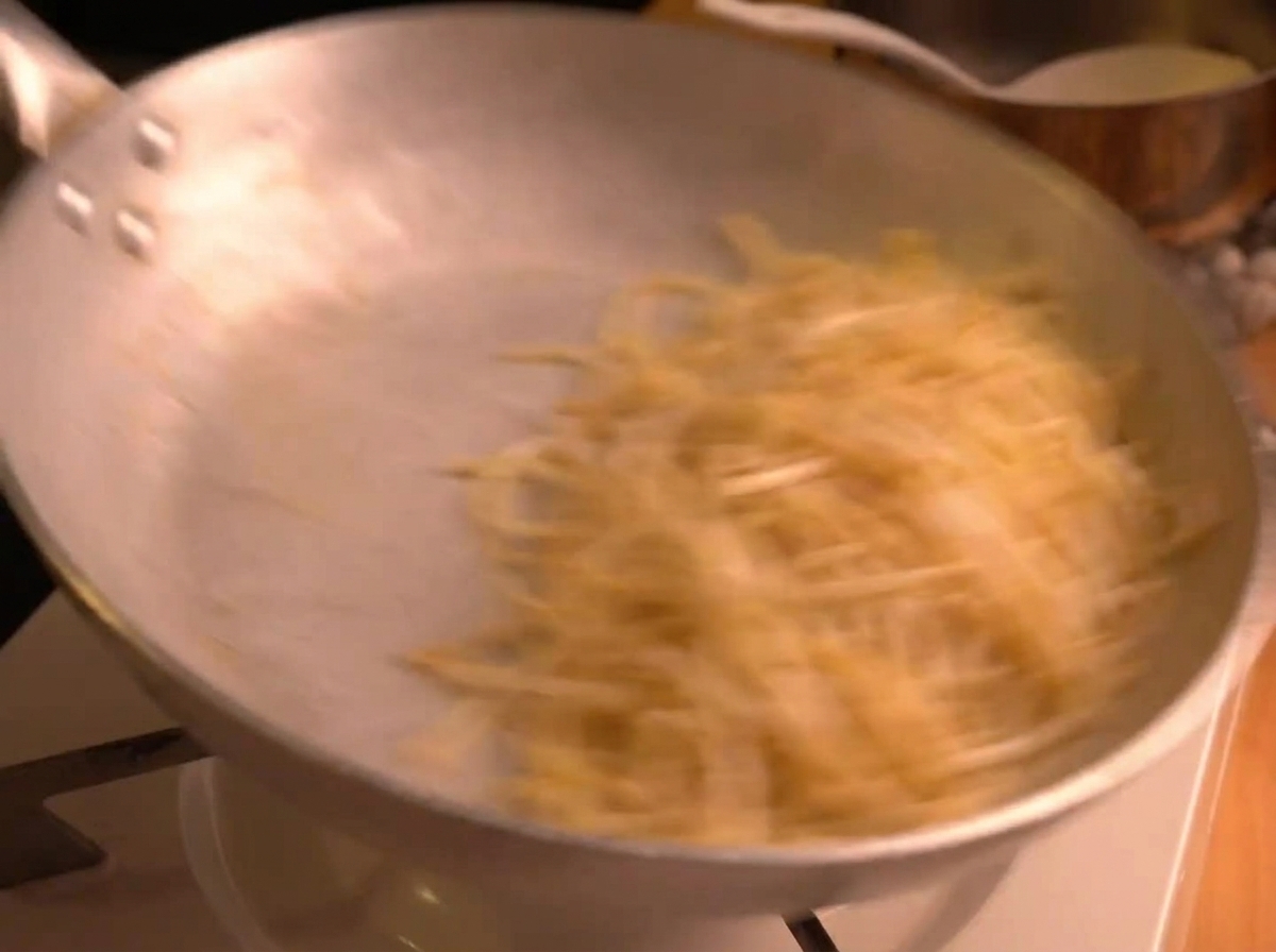 Spaghettis vigoureusement remués dans une poêle en inox, créant un flou de mouvement alors que la sauce sémulsionne.