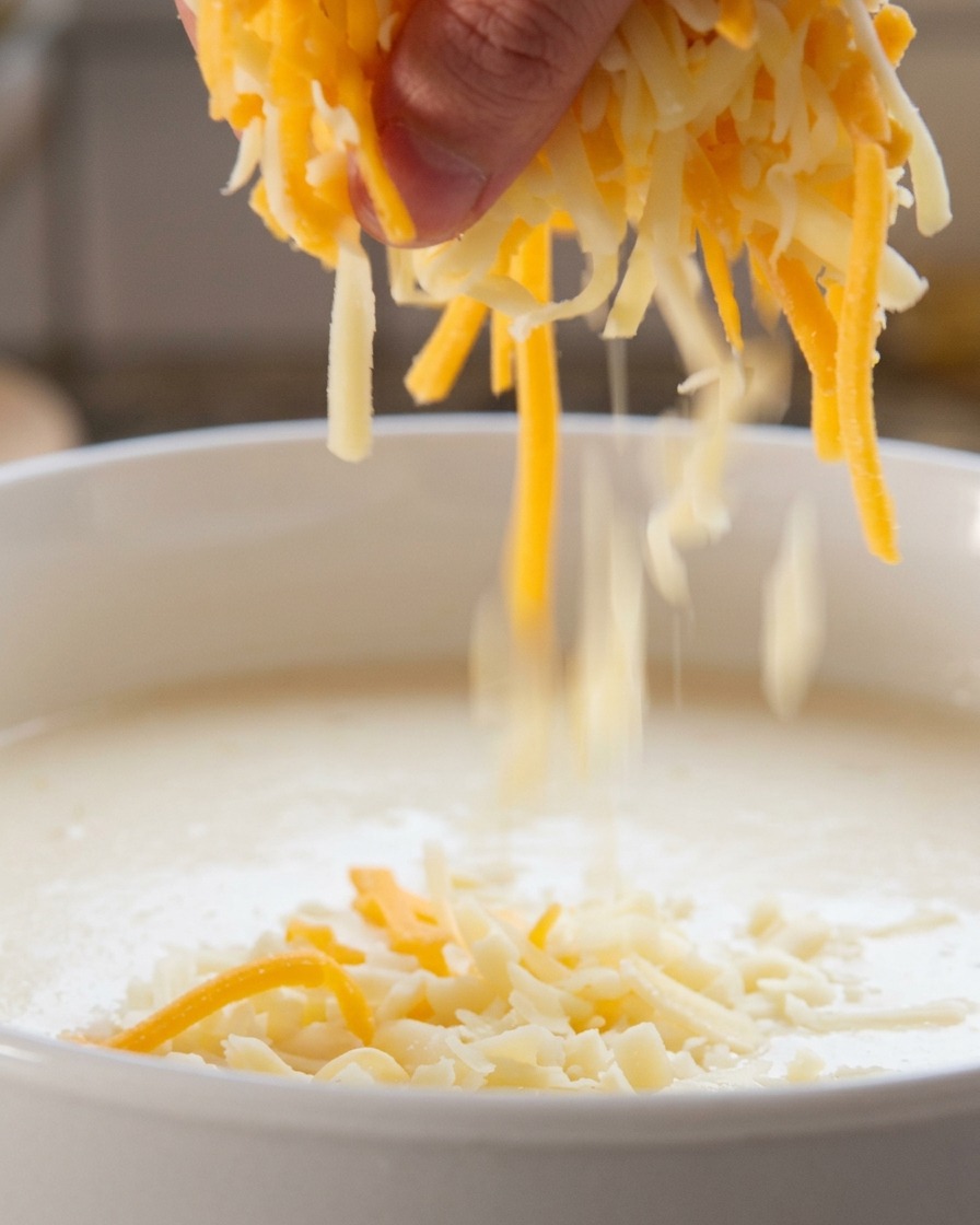 Une main saupoudrant un mélange de fromage blanc et orange râpé dans un liquide frémissant.