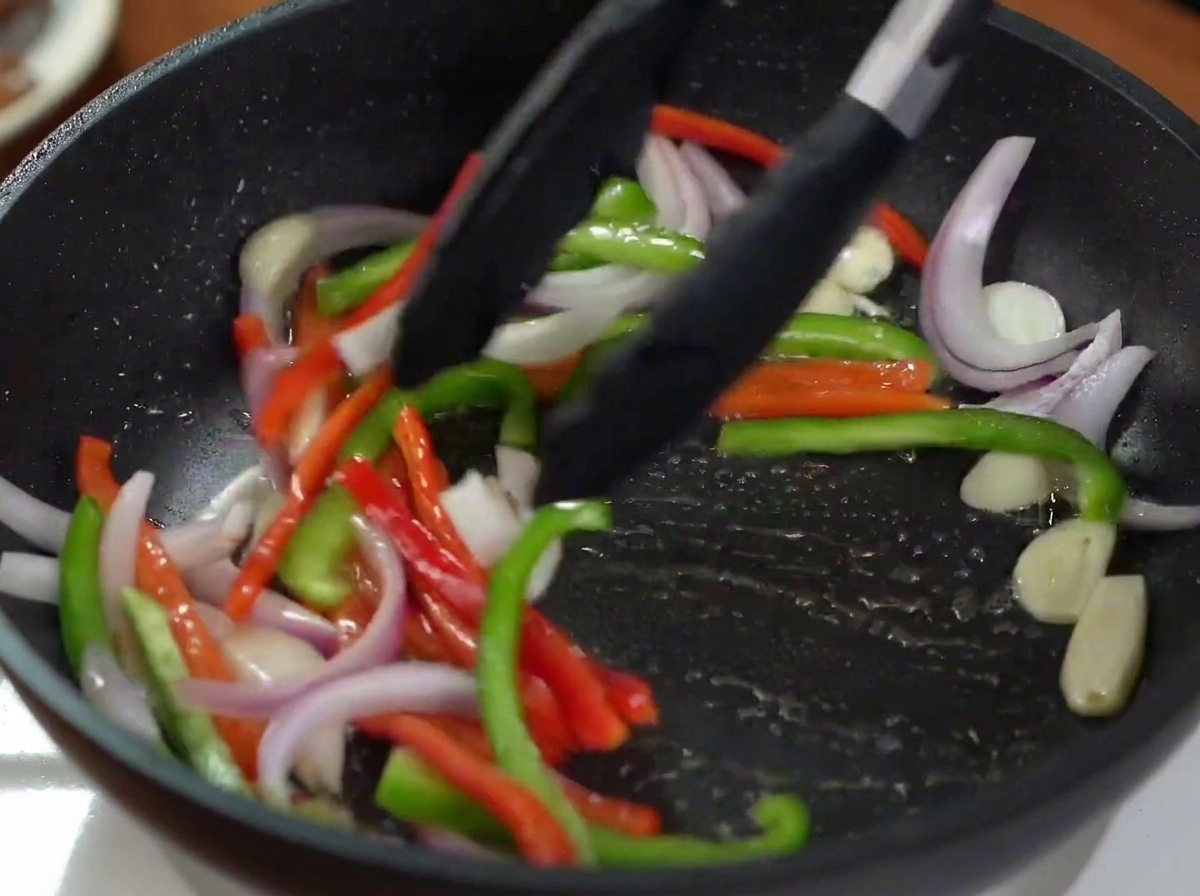 Pinces mélangeant des oignons rouges et des poivrons colorés dans une poêle chaude.