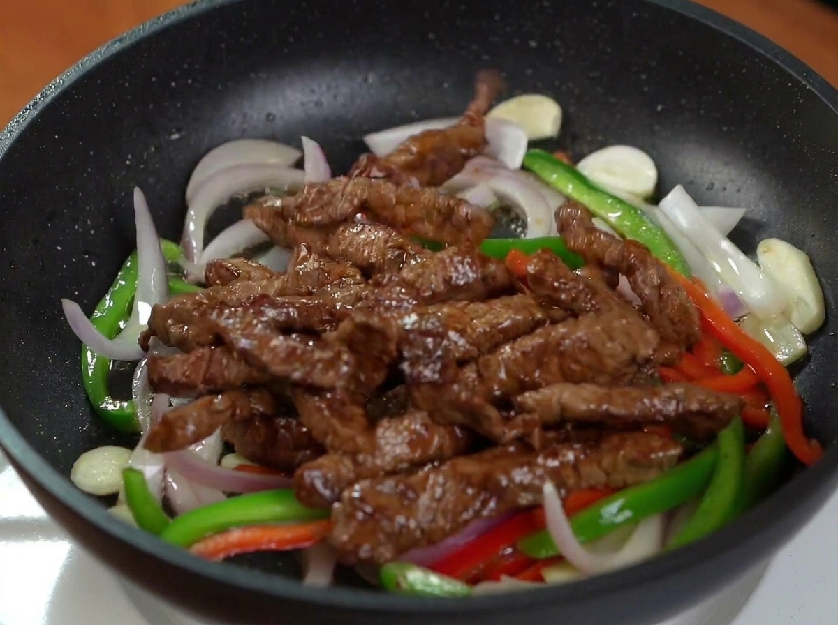 Lamelles de bœuf dorées ajoutées aux poivrons et oignons sautés dans une poêle noire.