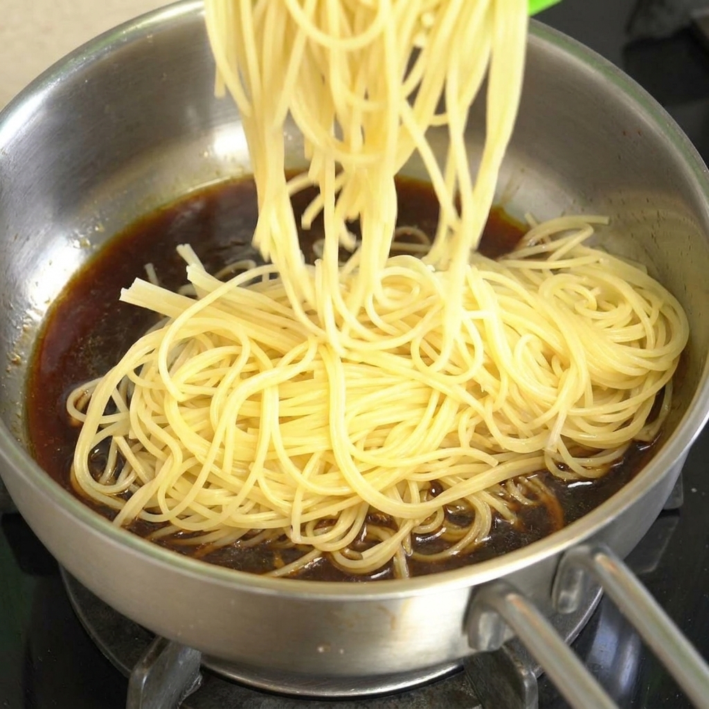 Spaghetti cuits ajoutés dans une poêle en acier inoxydable remplie de sauce brune au bœuf et poivre noir.