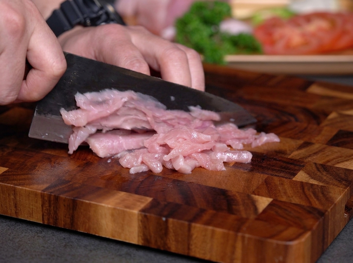 Mains tranchant de la viande maigre crue en fines lamelles sur une planche à découper avec un couteau sombre.