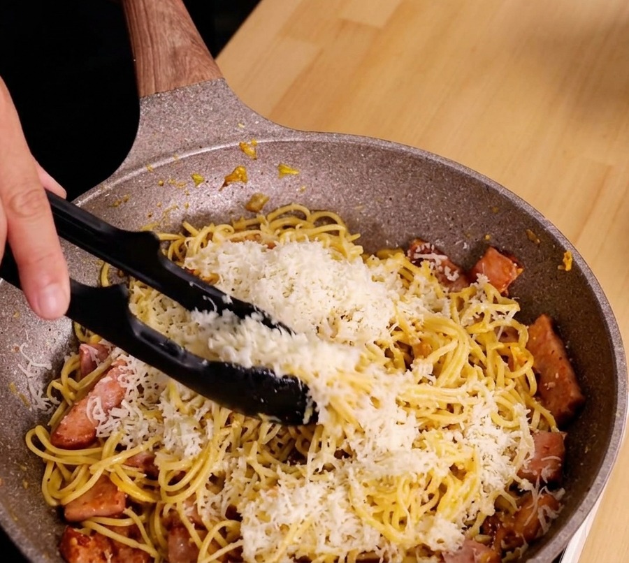 Mains utilisant une pince pour remuer activement les spaghettis, le fromage et le bacon dans une poêle afin de créer une sauce crémeuse.