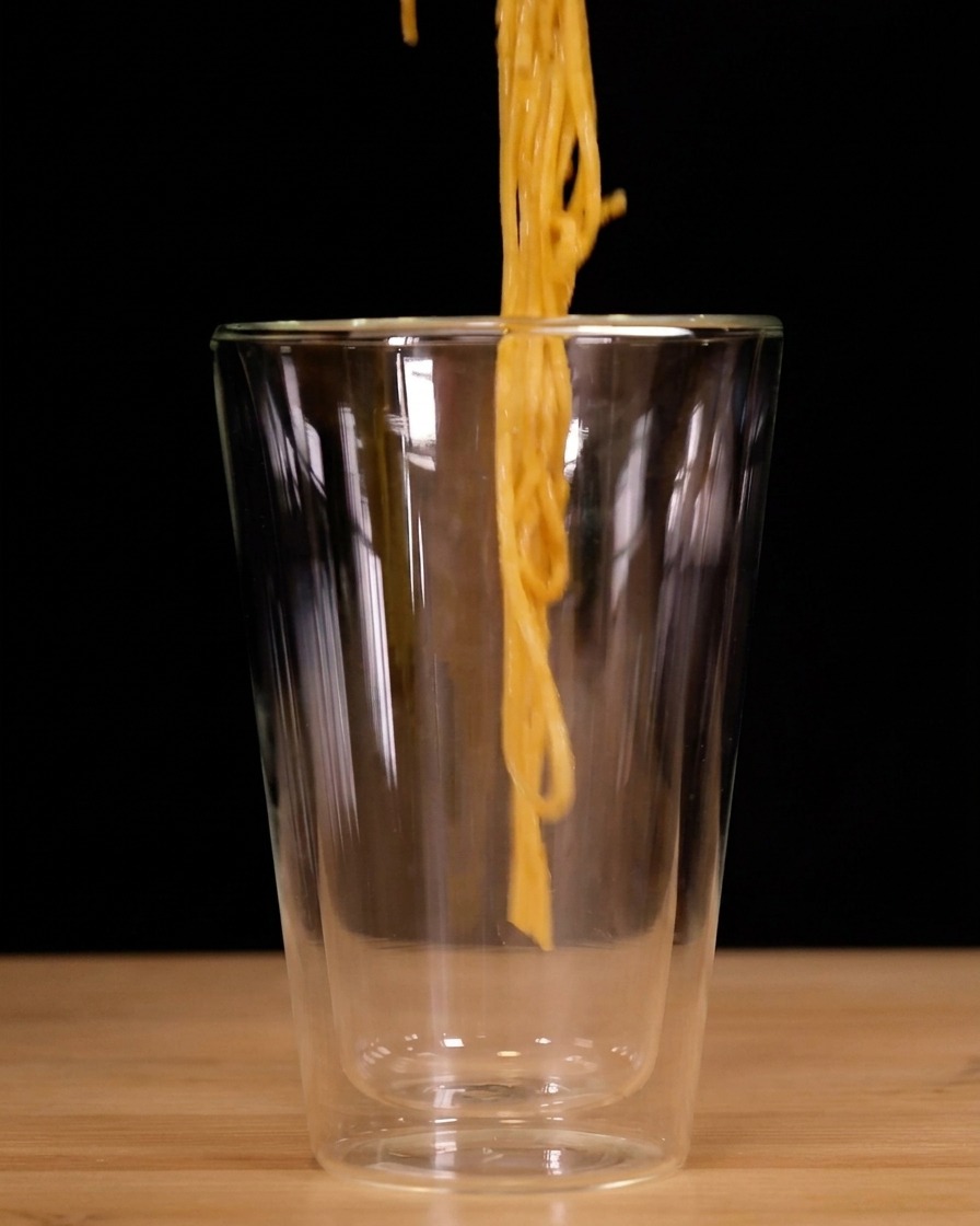 Spaghettis enroulés verticalement avec une pince et descendus dans un grand verre transparent à double paroi.