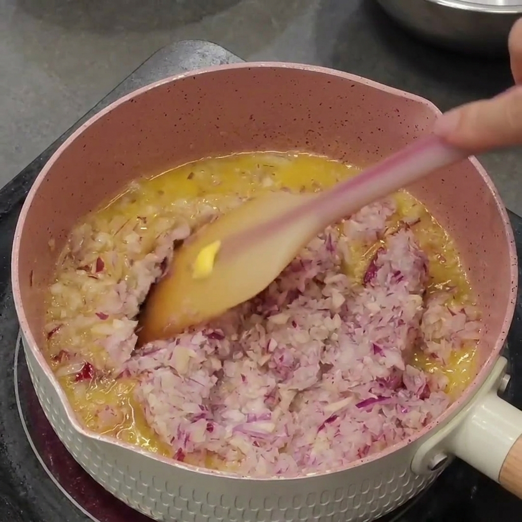 Oignons rouges et ail hachés revenant dans le beurre fondu à lintérieur dune casserole rose.