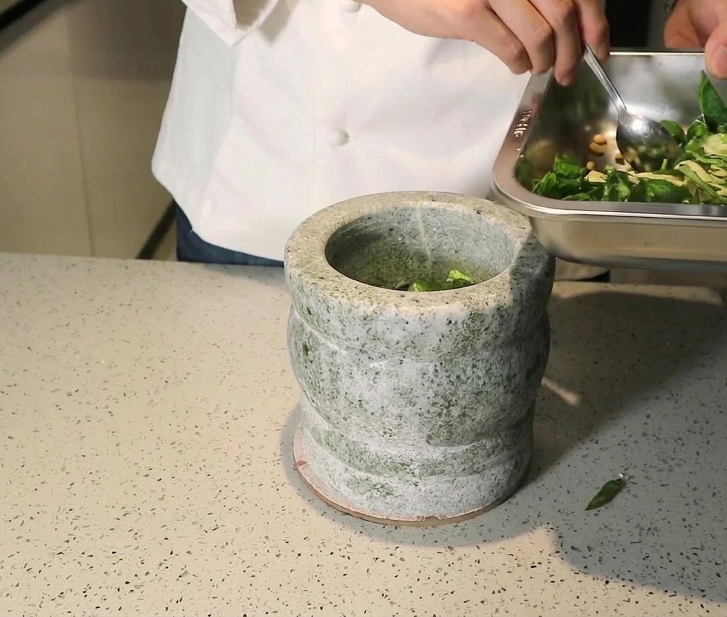 Chef transférant un mélange de basilic frais, pignons et ail dun plateau métallique vers un mortier en pierre.