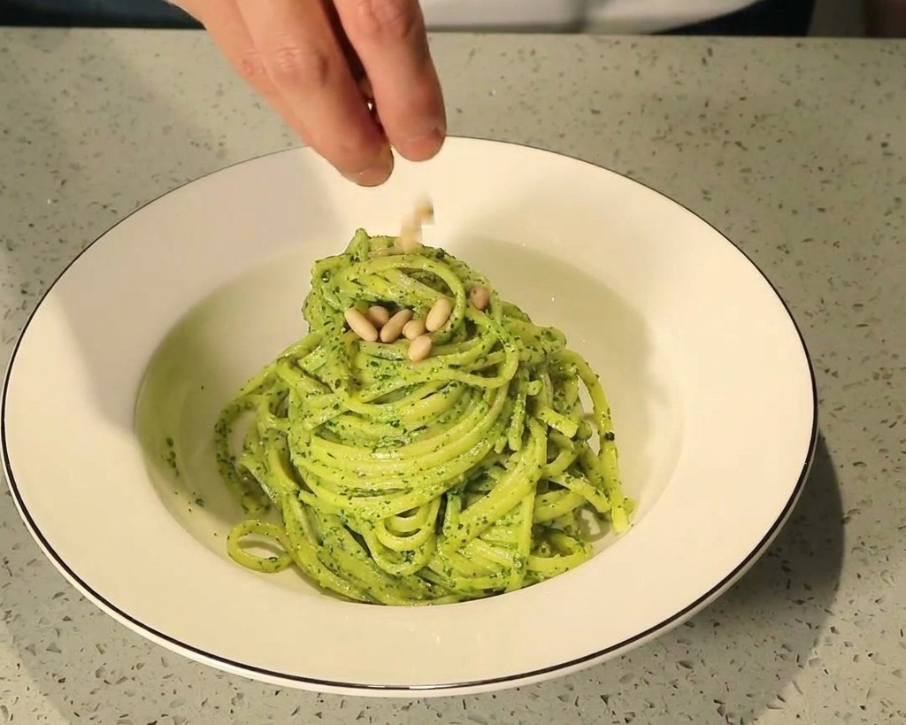 Doigts saupoudrant des pignons de pin grillés sur un dôme de pâtes au pesto dans un bol en céramique blanche.