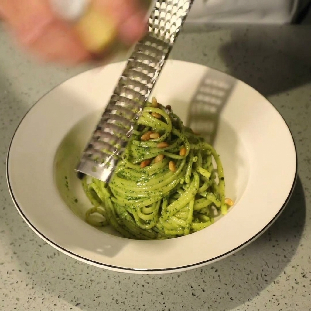 Une main utilisant une râpe pour ajouter du Parmesan frais sur un bol de pâtes au pesto garni de pignons.