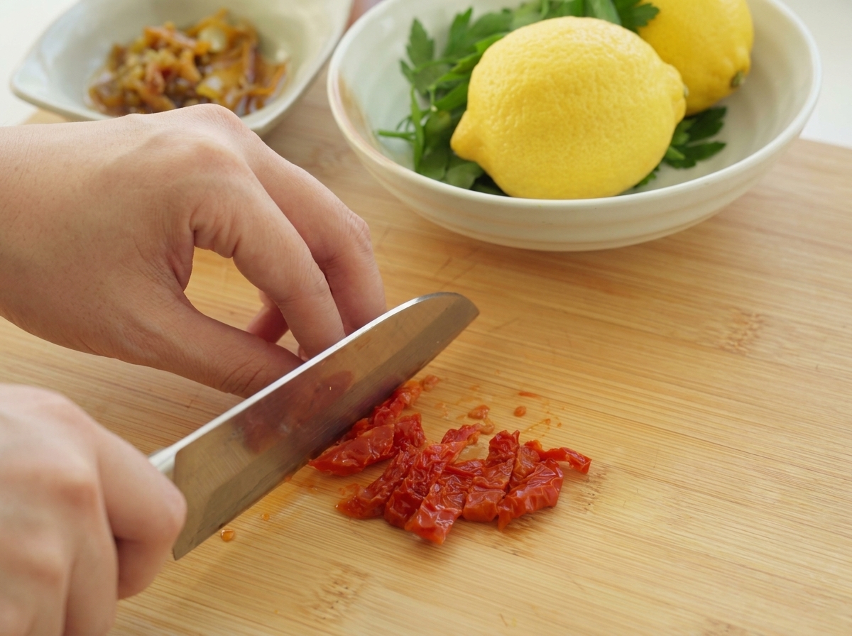 Une personne utilisant un couteau de chef pour hacher finement des tomates séchées rouge vif sur une planche en bois.