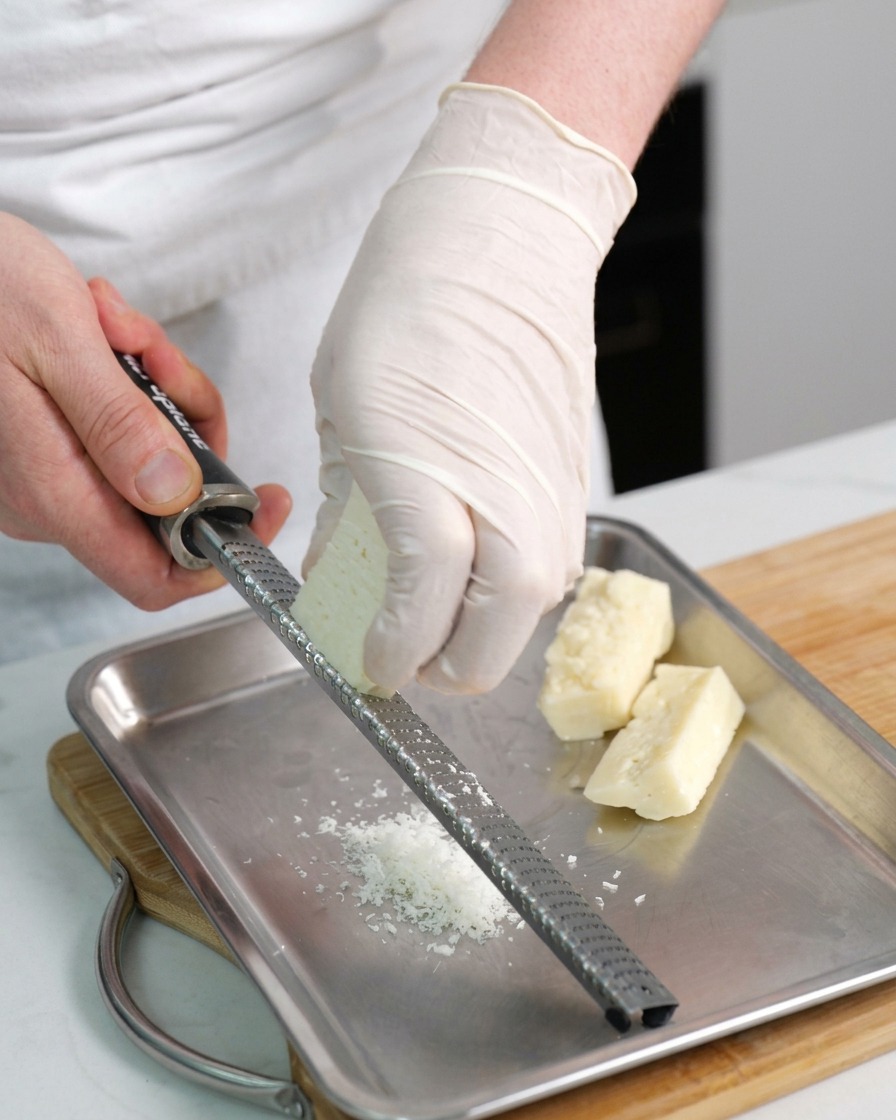 Mains gantées utilisant une râpe pour transformer un bloc de Pecorino en fins copeaux sur un plateau.