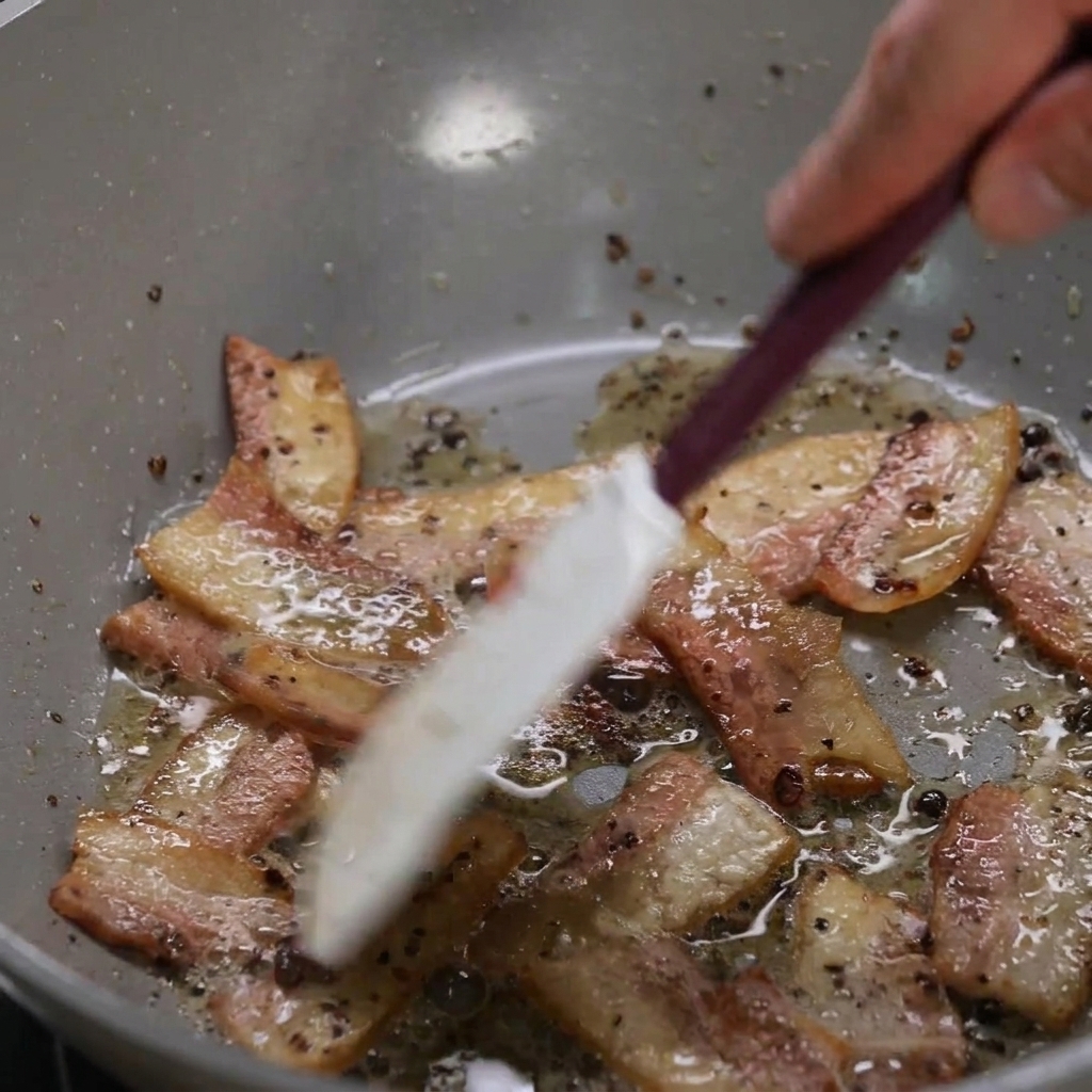 Lanières de guanciale dorant dans une poêle, rendant leur gras translucide sous laction dune spatule.