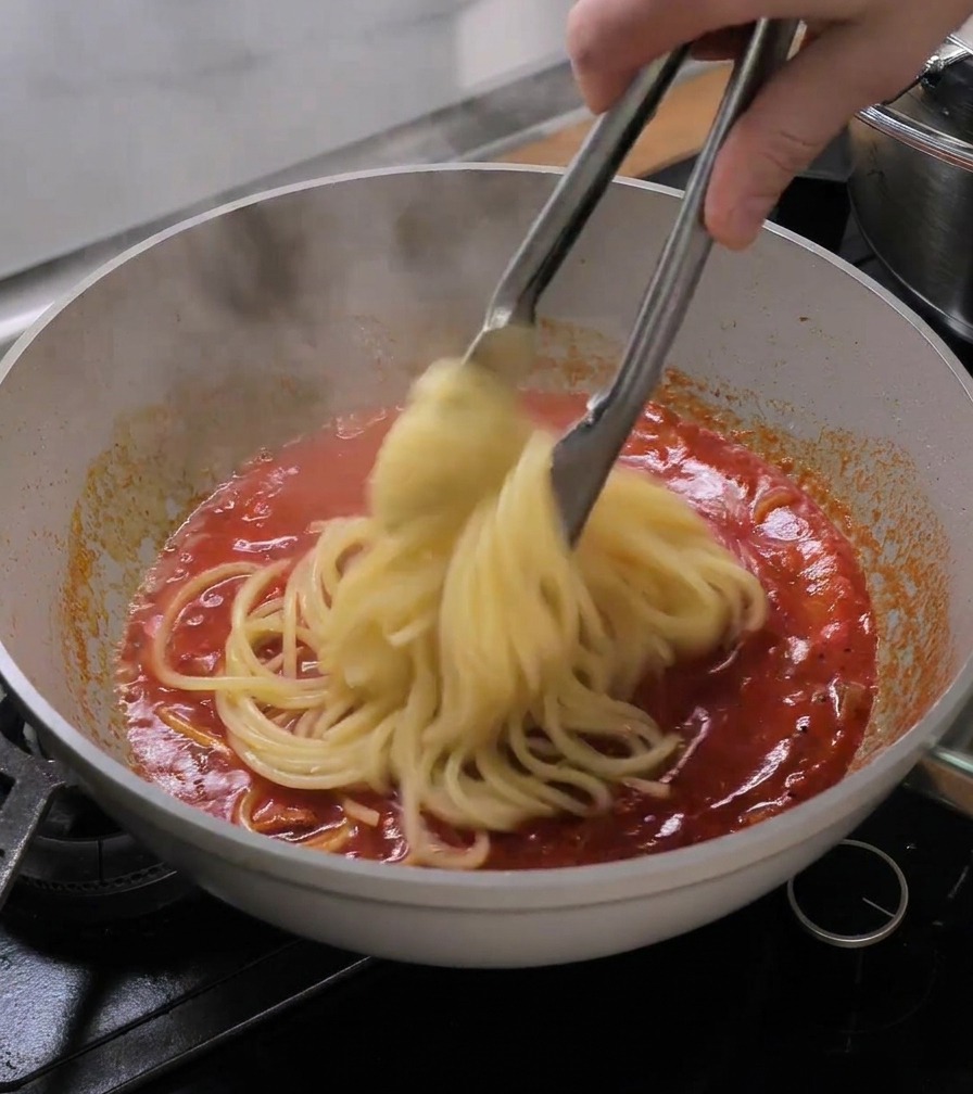 Pince de cuisine transférant les bucatini directement de leau vers la poêle de sauce tomate fumante.