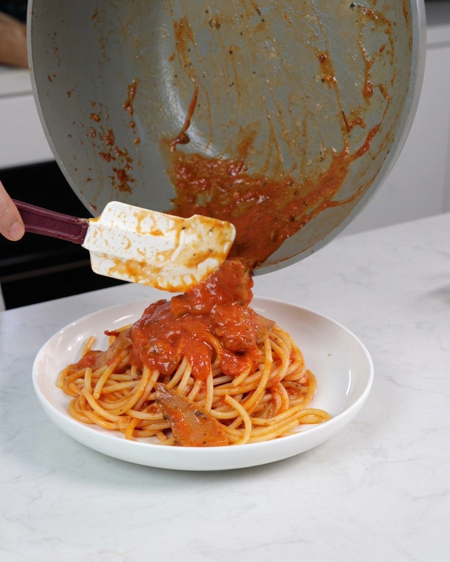 Utilisation dune spatule pour verser le reste de sauce tomate et de morceaux de viande sur les pâtes dans un bol de service.
