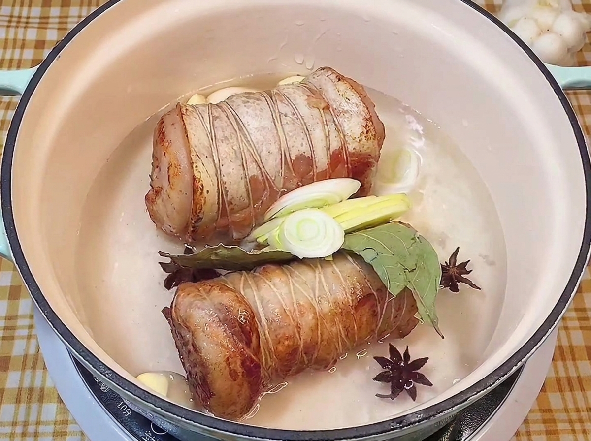 Rouleaux de porc dorés reposant dans une marmite avec badiane, laurier, ail et poireaux frais.
