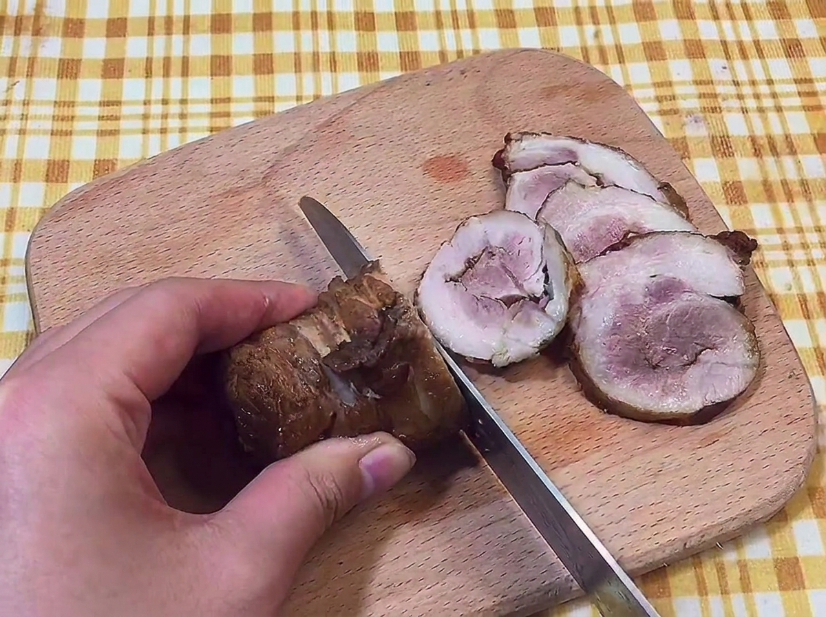 Main tenant un couteau pour couper un rouleau de porc braisé en tranches fines sur une planche en bois.