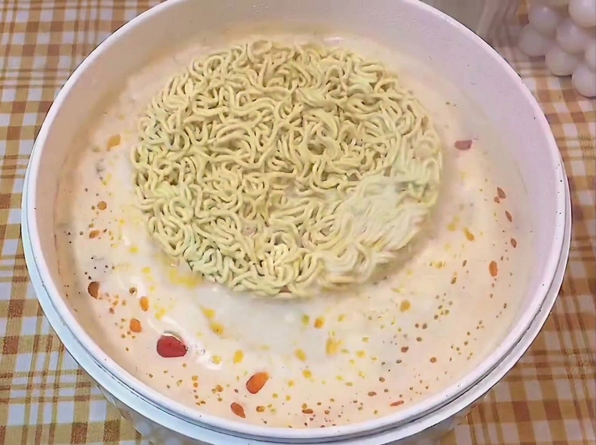 Un bloc rond de nouilles ramen sèches flottant dans une casserole blanche remplie de bouillon assaisonné.