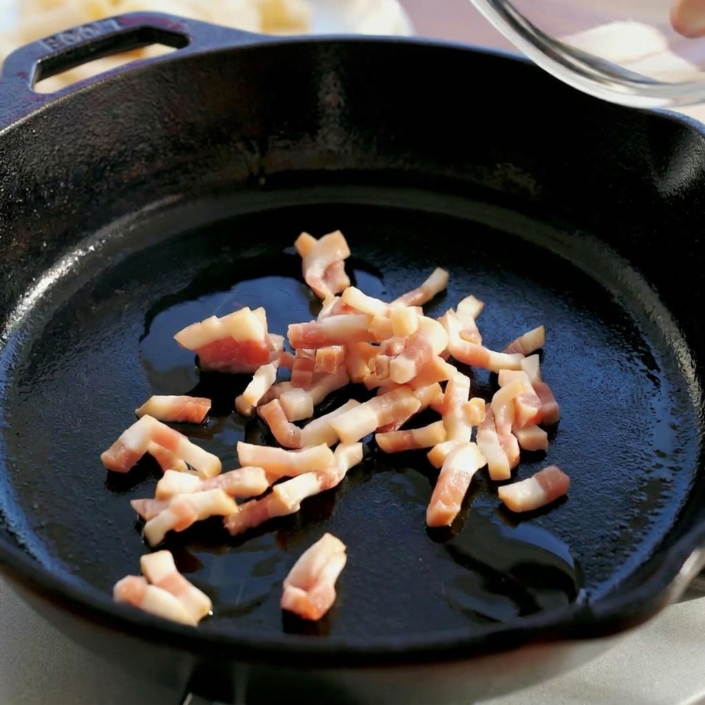 Du guanciale cru en dés versé depuis un bol en verre dans une poêle en fonte noire.