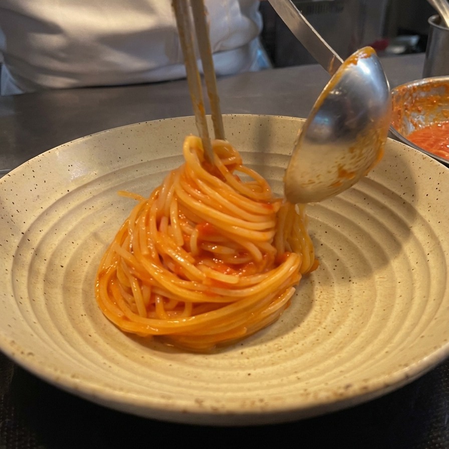 Un chef utilisant des pinces et une louche en métal pour enrouler des spaghettis à la tomate en un dôme soigné sur une assiette en céramique rustique.