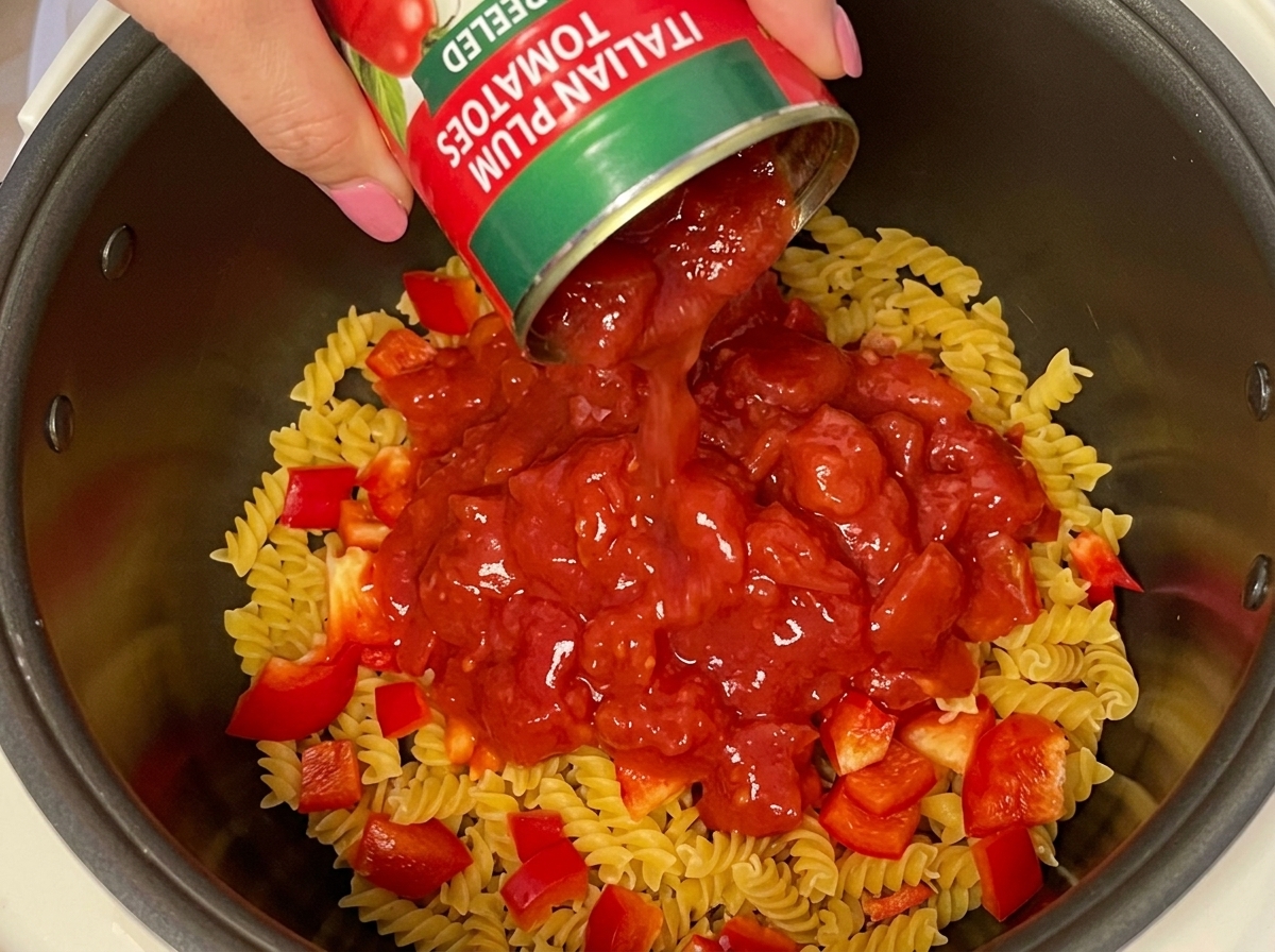Une main versant une conserve de tomates concassées sur les pâtes et les poivrons dans le cuiseur à riz.