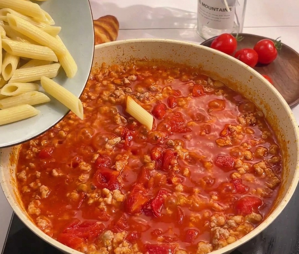 Des pennes bouillies versées dun bol dans une poêle frémissante de sauce à la tomate et à la viande.