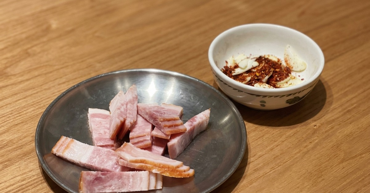Bacon cru tranché sur une assiette à côté dun bol dail émincé et de flocons de piment sur une table en bois.