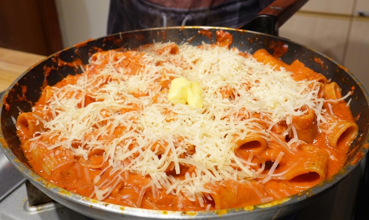 Un cube de beurre jaune posé sur une pile de parmesan râpé et de rigatoni dans une poêle chaude.