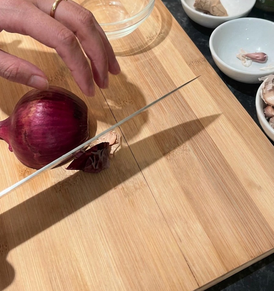 Une main tenant un oignon rouge sur une planche à découper en bois avec un couteau de chef prêt à trancher.