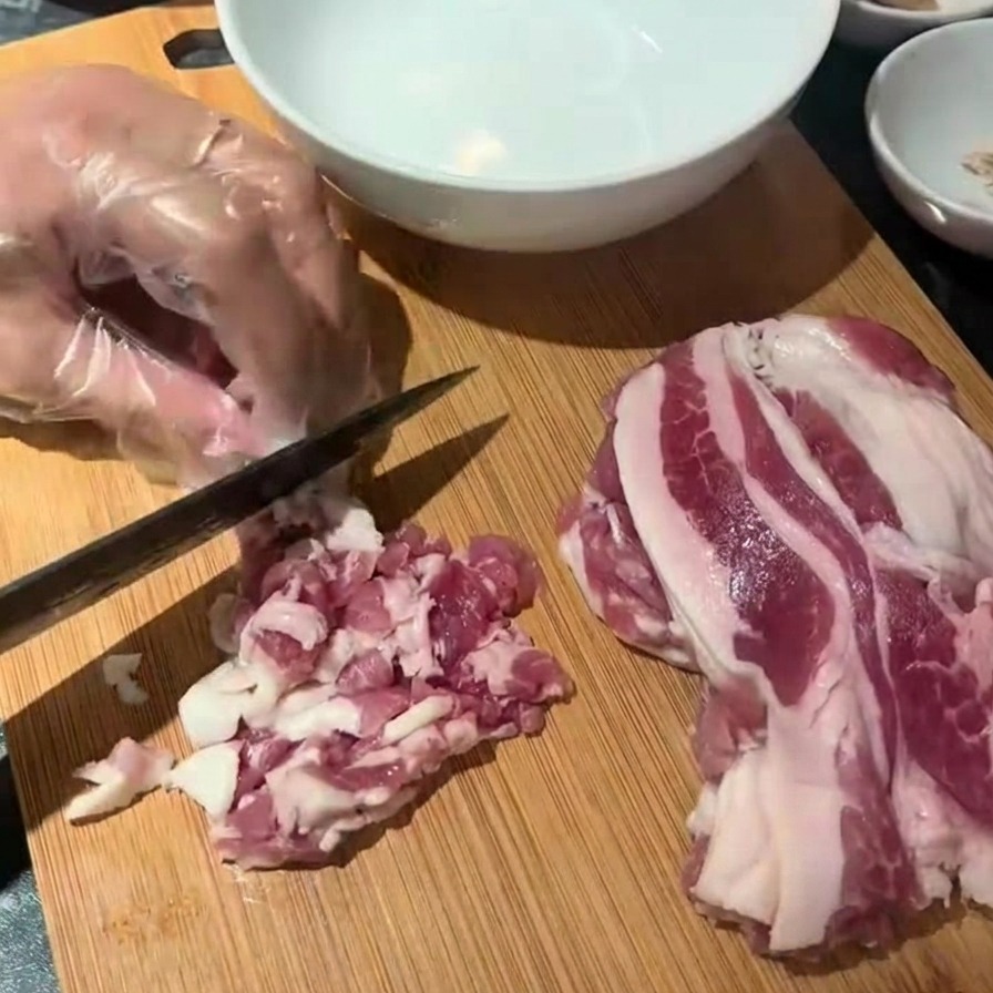 Une main gantée utilisant un couteau noir pour couper des tranches de bacon cru sur une planche à découper.