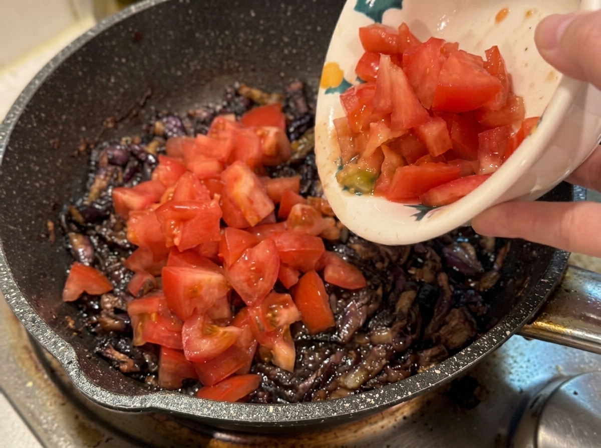 Une main versant des tomates fraîches en dés dans une poêle chaude contenant du bacon et des oignons.