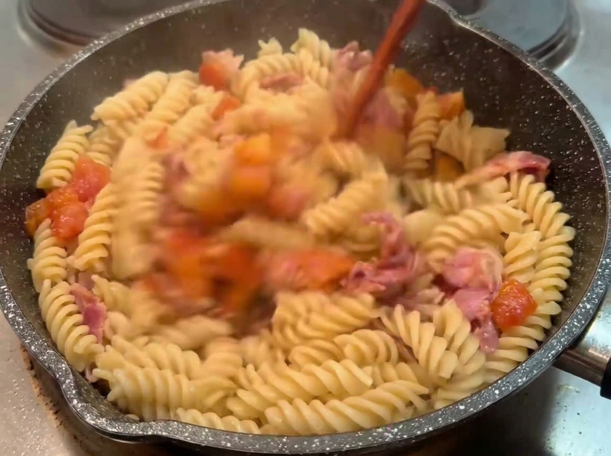 Fusilli cuits mélangés dans une poêle sombre avec une sauce tomate et bacon généreuse à laide dun ustensile en bois.